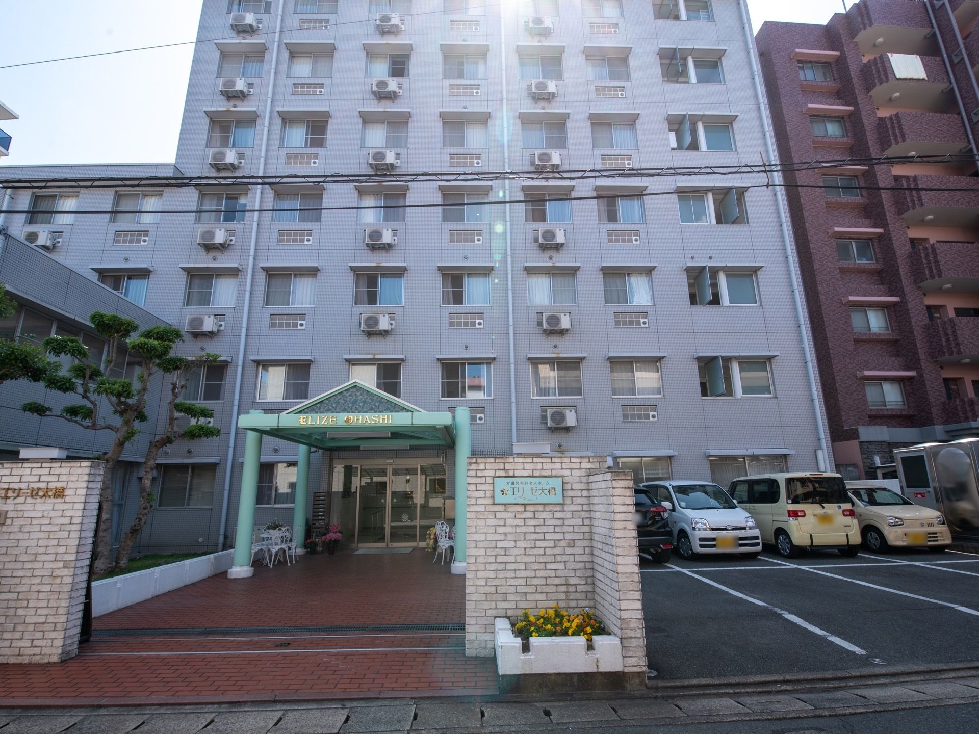 道路に面した施設入口。両側にレンガ積みの門柱が建っている。右側の柱には、薄い緑色の看板が設置されている。