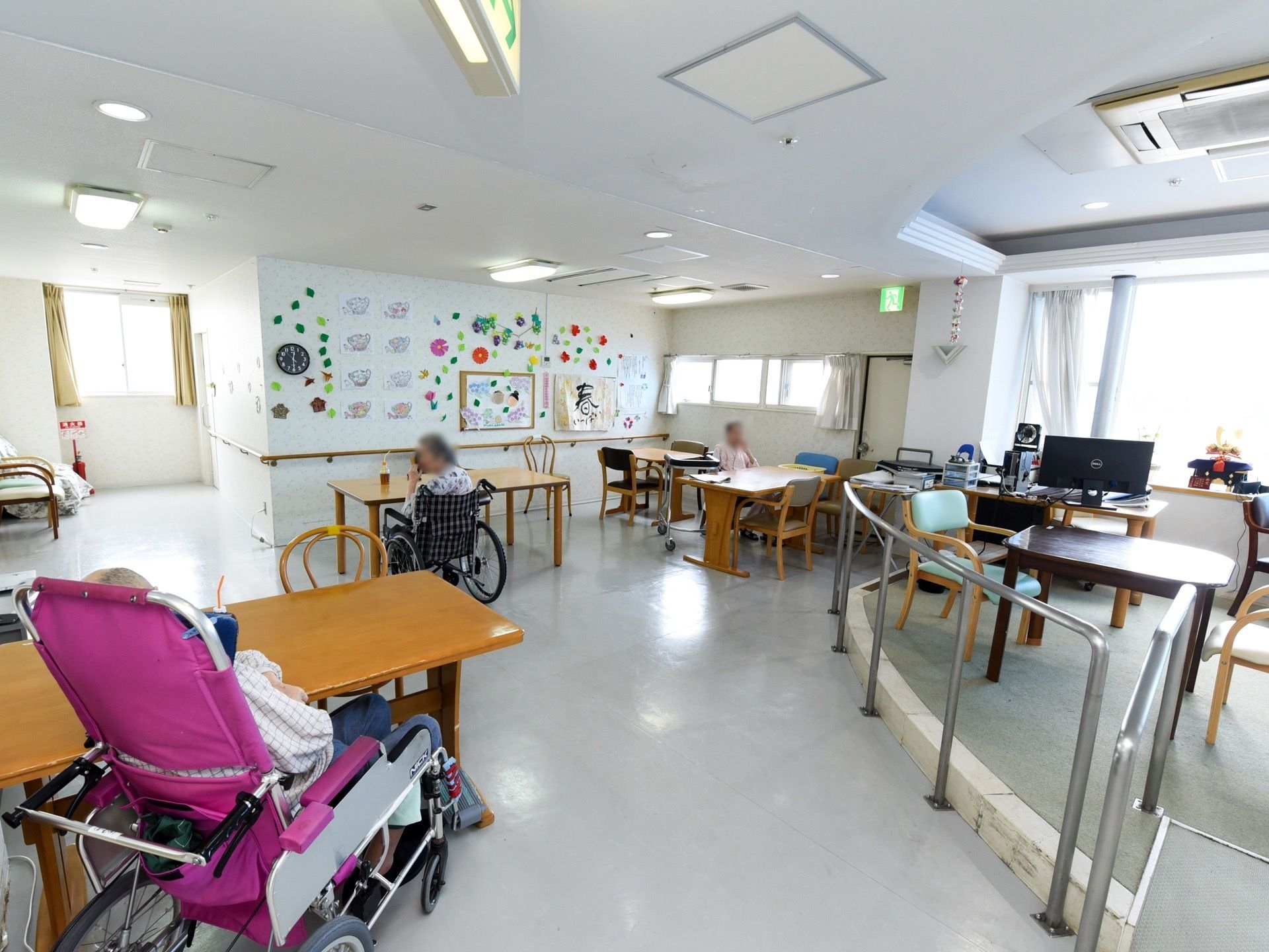 長方形のテーブルが複数台置かれている。車椅子をご利用の方々が思い思いにくつろいでいる。壁沿いやスペース中央部には手すりが備わっている。