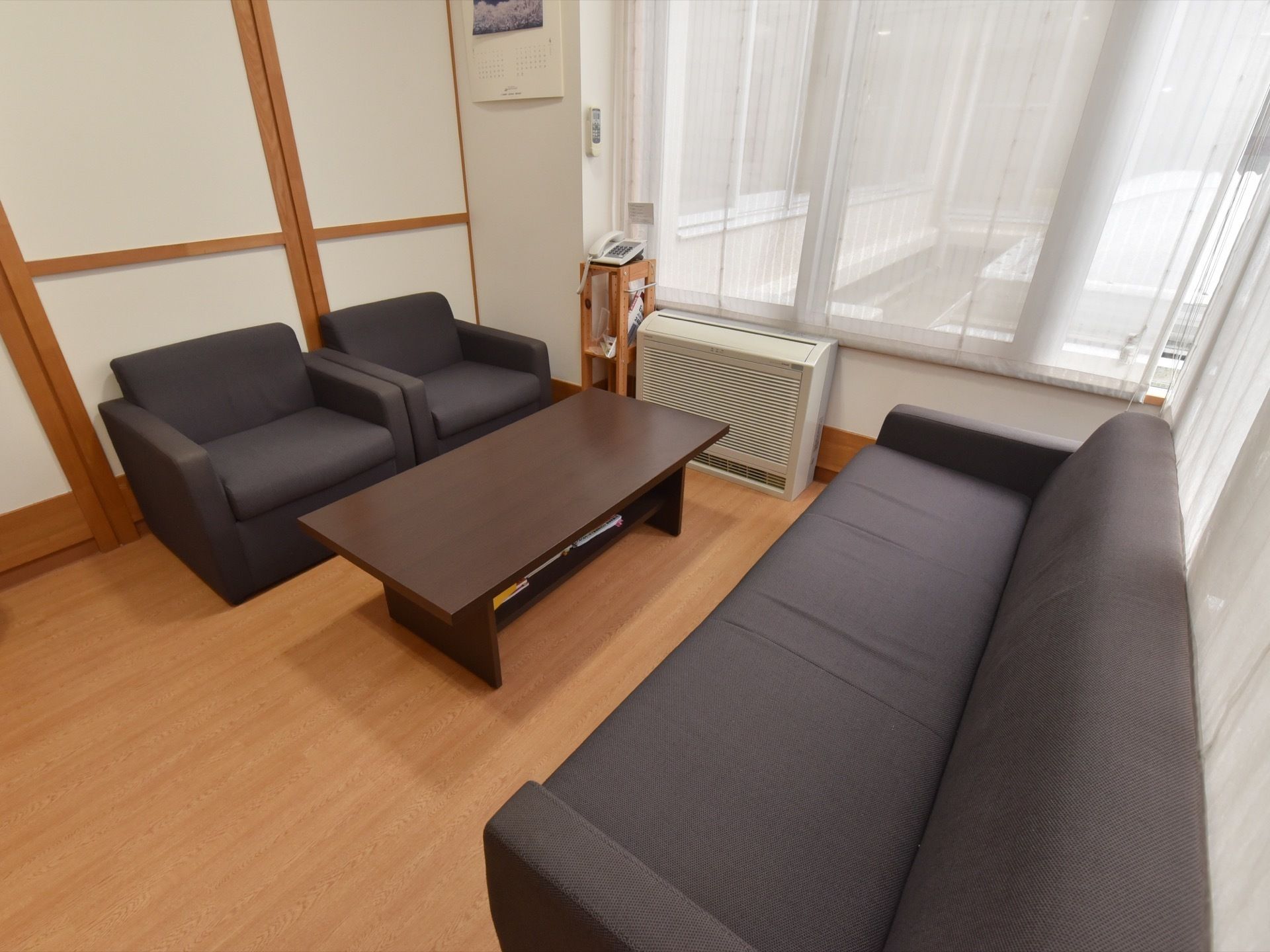 大きな窓が２面にある明るい空間にゆったり座れる落ち着いた色の応接セットが設置されている。棚の上に電話機が置かれている。