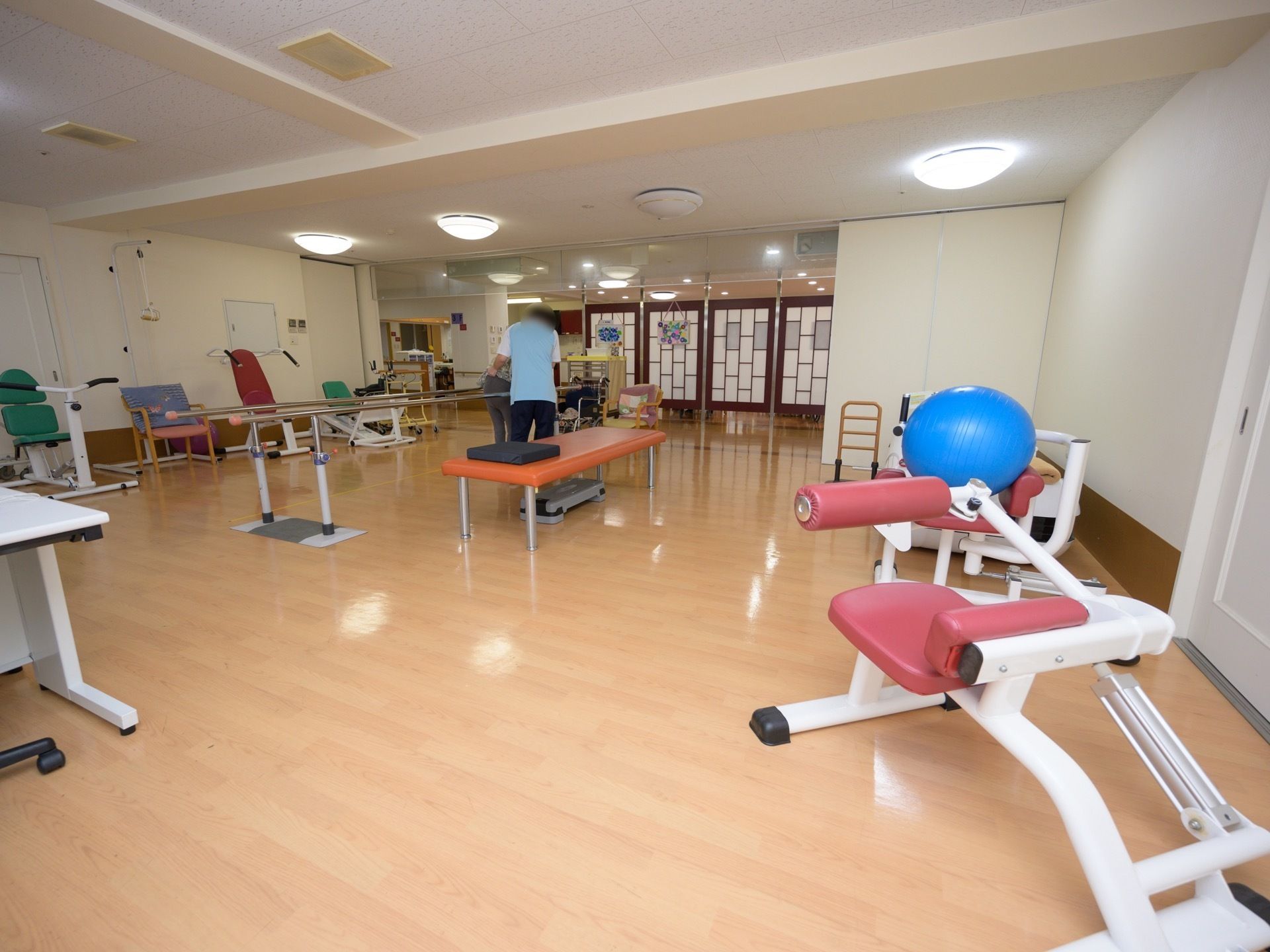 ベージュ色の床に白色でまとめられた、広い空間の部屋である。色々な種類のリハビリ用の器具が並んでいる。