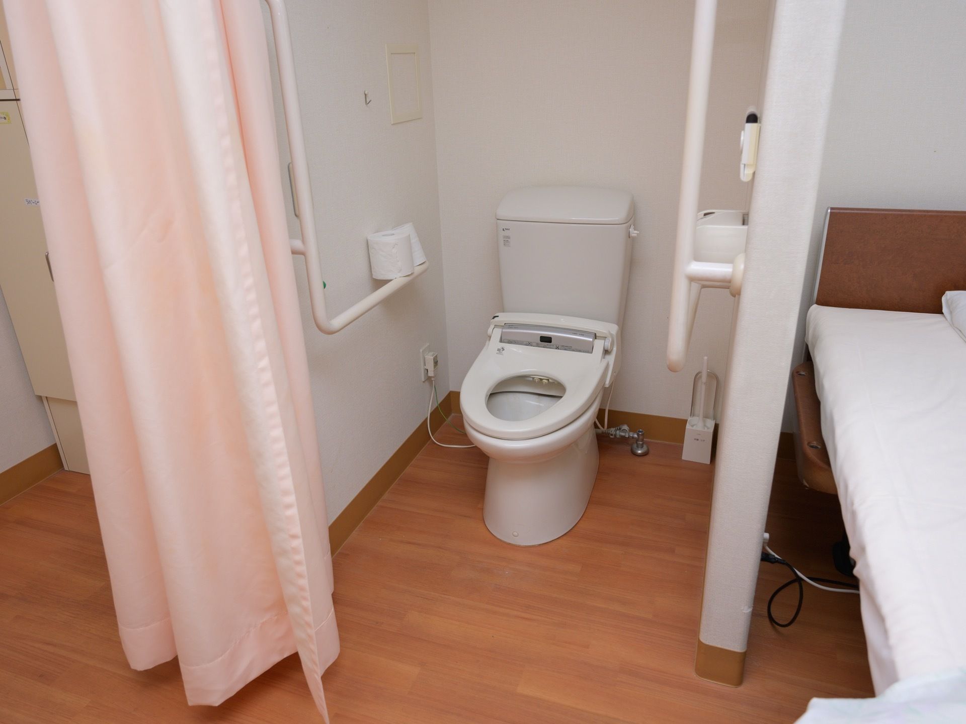 居室の中にトイレがある。カーテンで仕切られており、白い温水洗浄便座の便器がある。壁にＬ字の手すりがある。