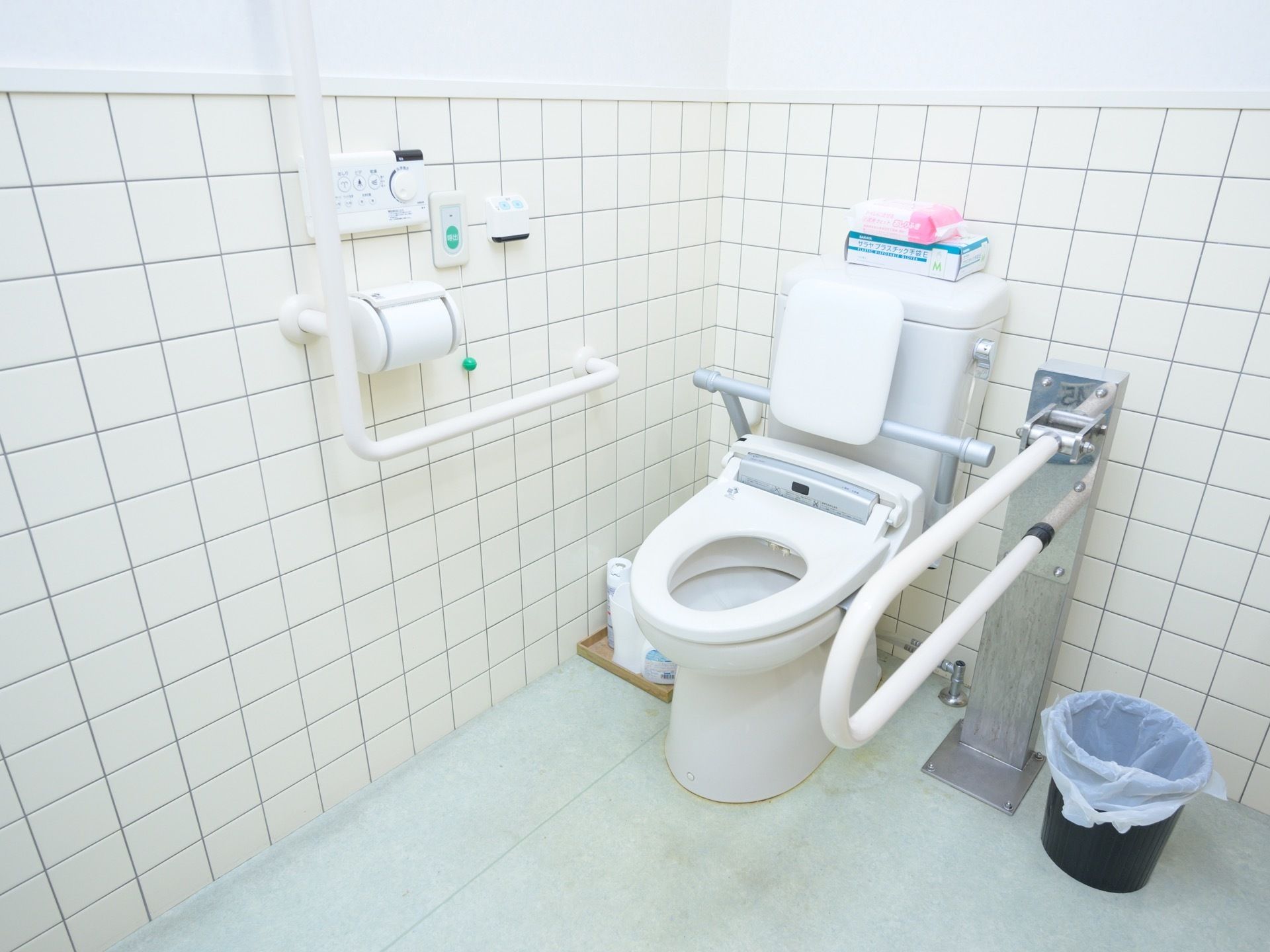 壁がタイル貼りのトイレである。温水洗浄便座にクッション性のある背もたれがついている。可動式の手すりがある。