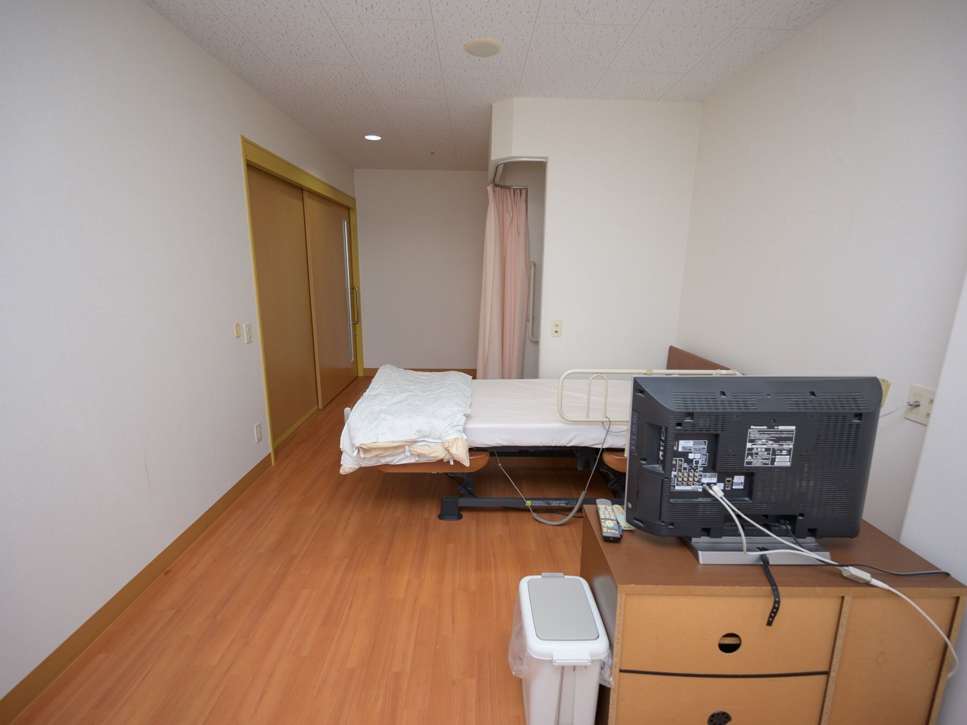 ベッドとテレビが備え付けられた居室である。壁には引き扉と仕切り用のカーテンがある。テレビは収納ボックスの上に乗っている。