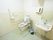温水洗浄便座が奥にあり、隣に洗面台があるトイレスペースである。便座に可動式の手すりがある。洗面台は横長の四角いものである。