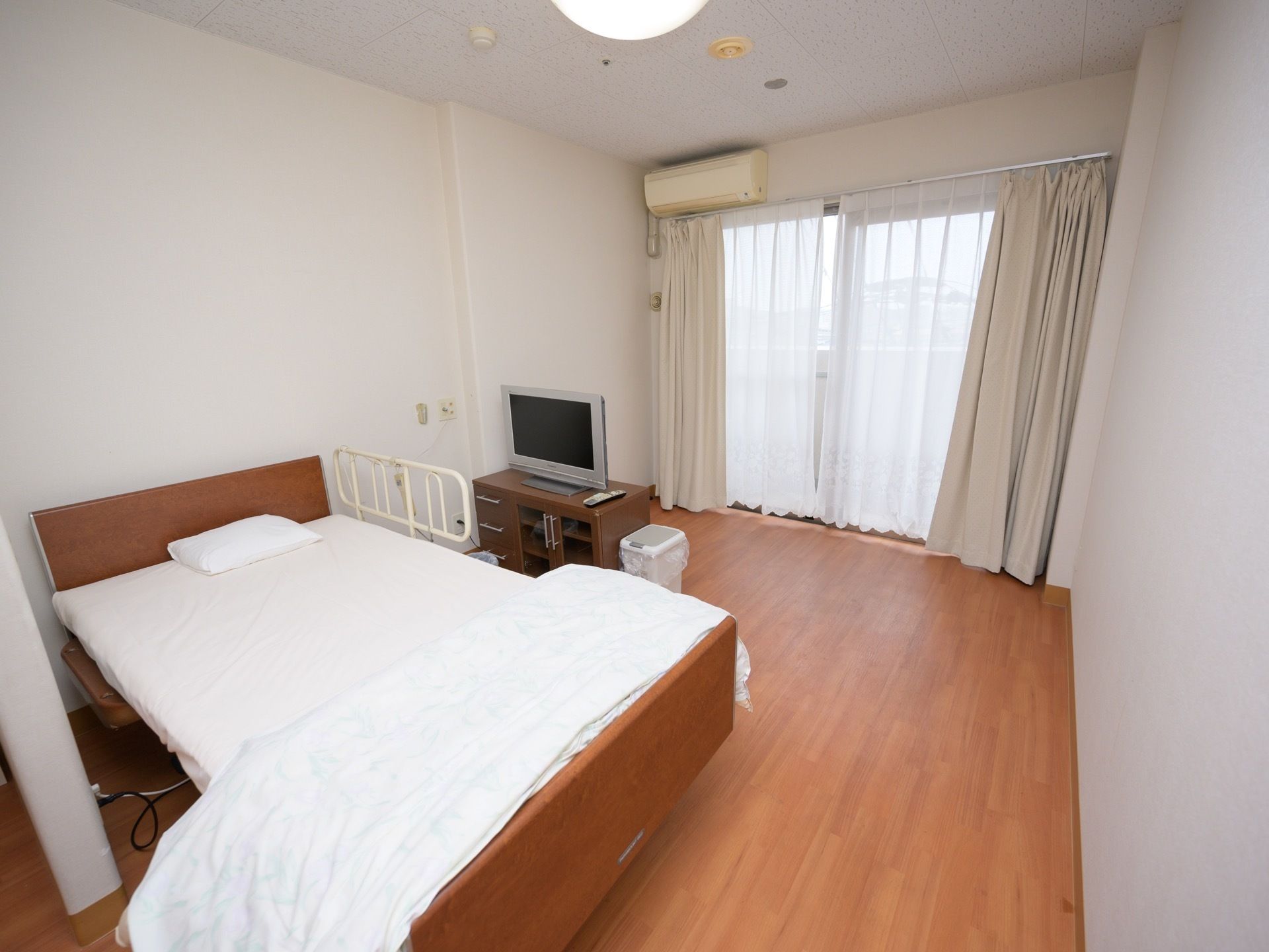突き当りに掃出し窓があり、カーテンが掛かっている。窓から少し控えた所にテレビがあり、その前にベッドがある。