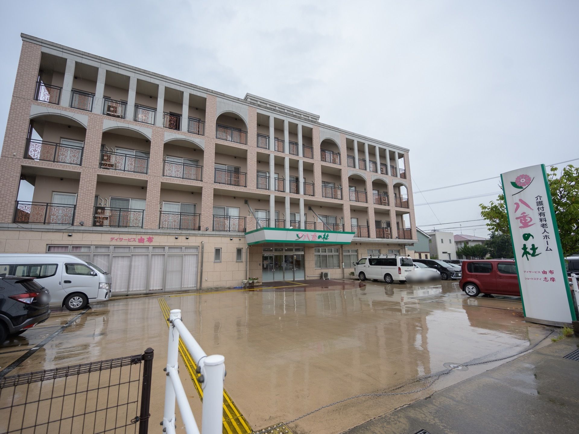 ４階建ての施設であり、施設名板に介護付有料老人ホームと明記されている。玄関前まで点字ブロックが続いている。建物前には駐車場が広がっている。
