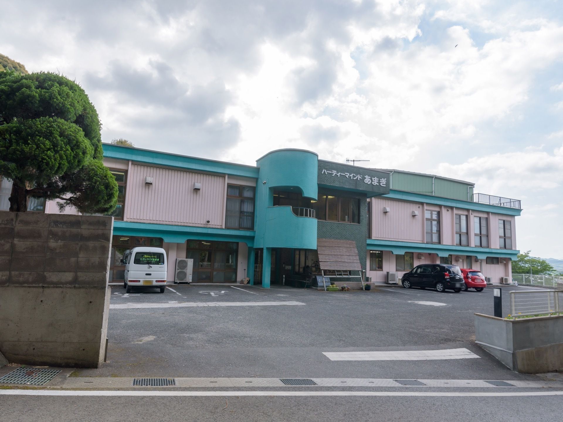 道路からすぐの場所に建っている２階建ての施設である。ピンクと緑のツートンカラーの外壁が特徴的。建物の前に車が３台停まっている。