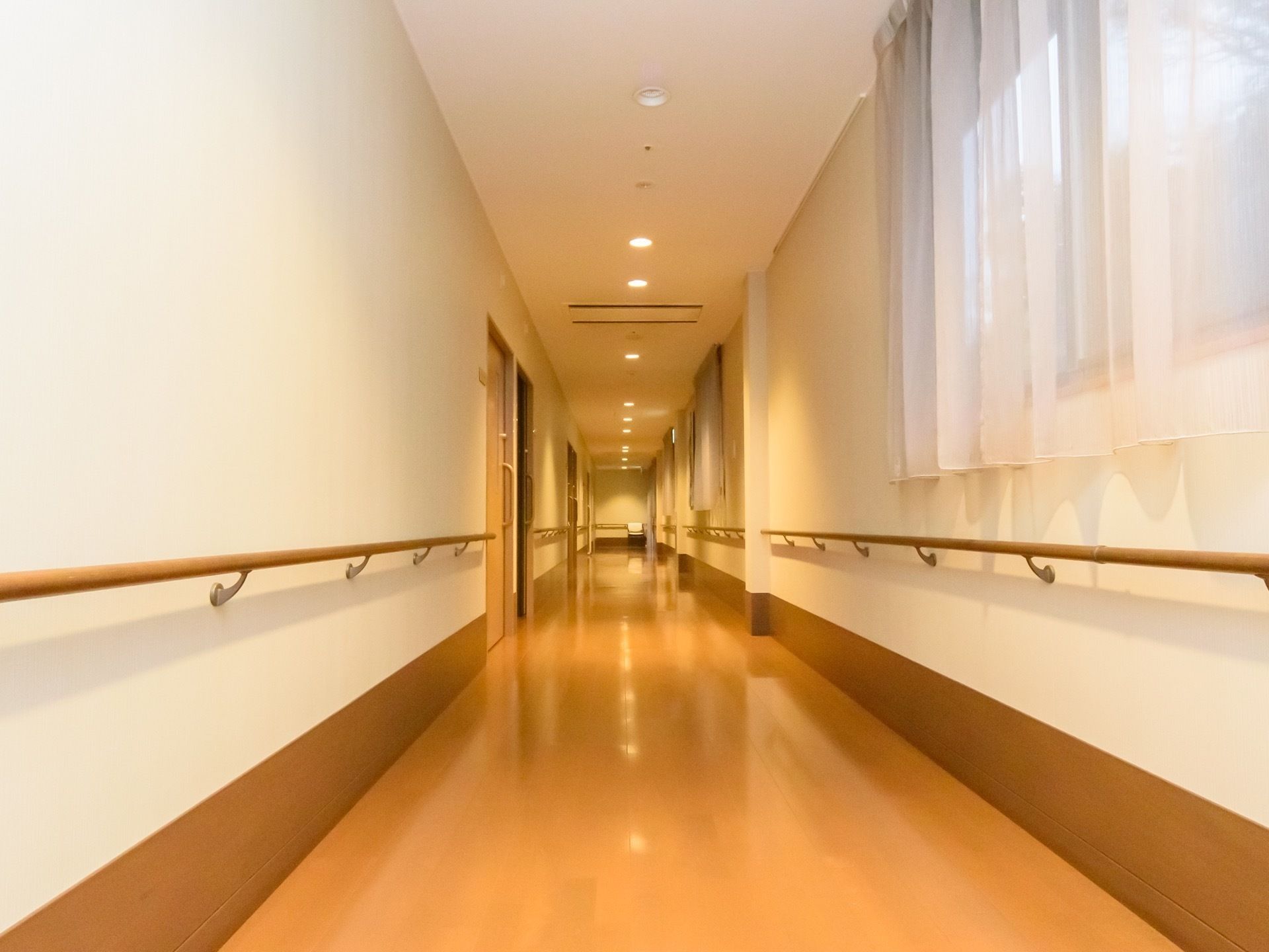壁に設置してある手すりが入居者様の歩行をサポートしている。白と茶の配色にオレンジ系の照明が目に優しい。真っ直ぐな通路は、安全に歩行できる。