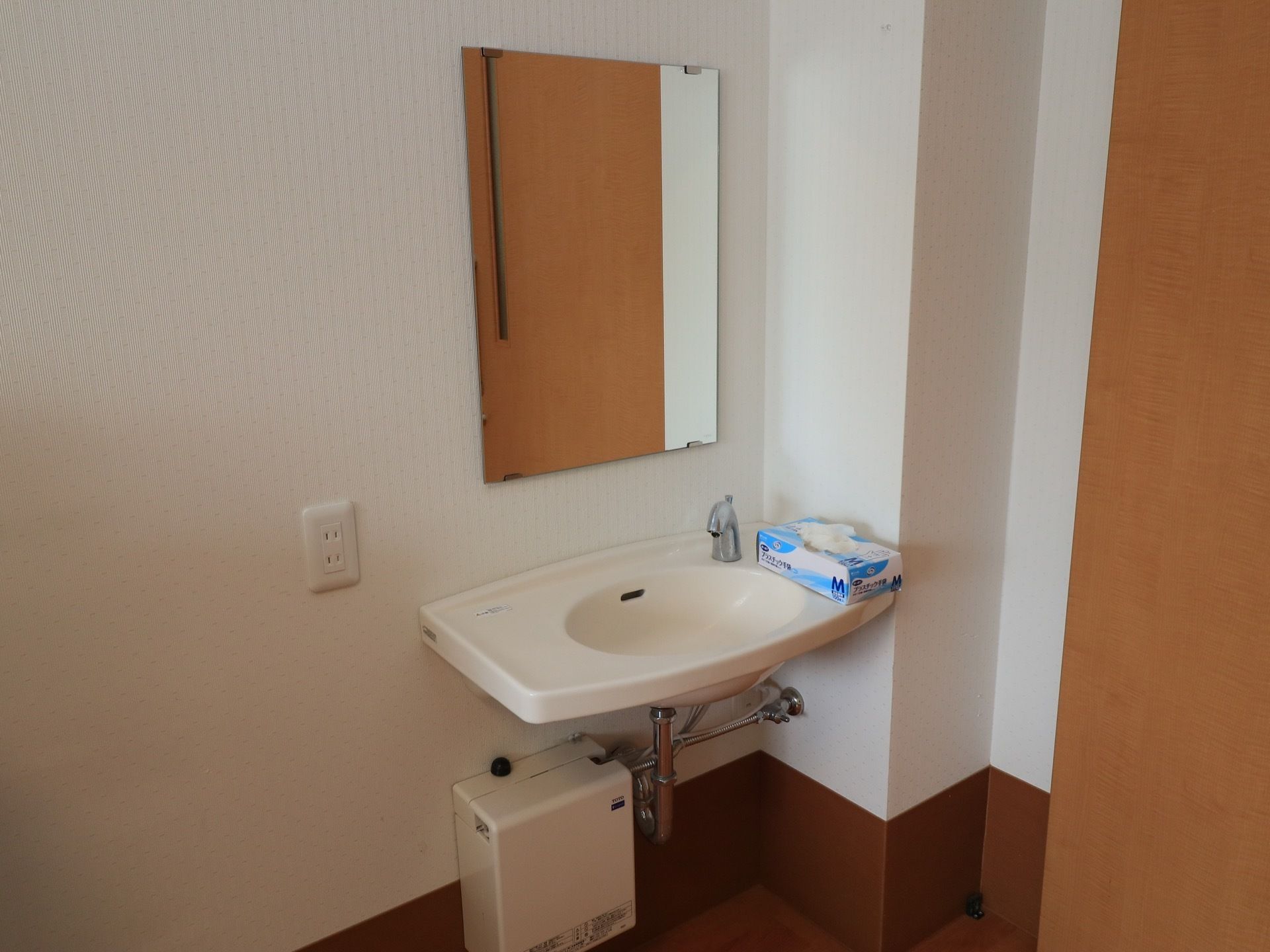 部屋の角に洗面台がある。壁に直接ボウルがかけられていおり水栓は自動である。壁に鏡が貼られ、そばにコンセントがある。