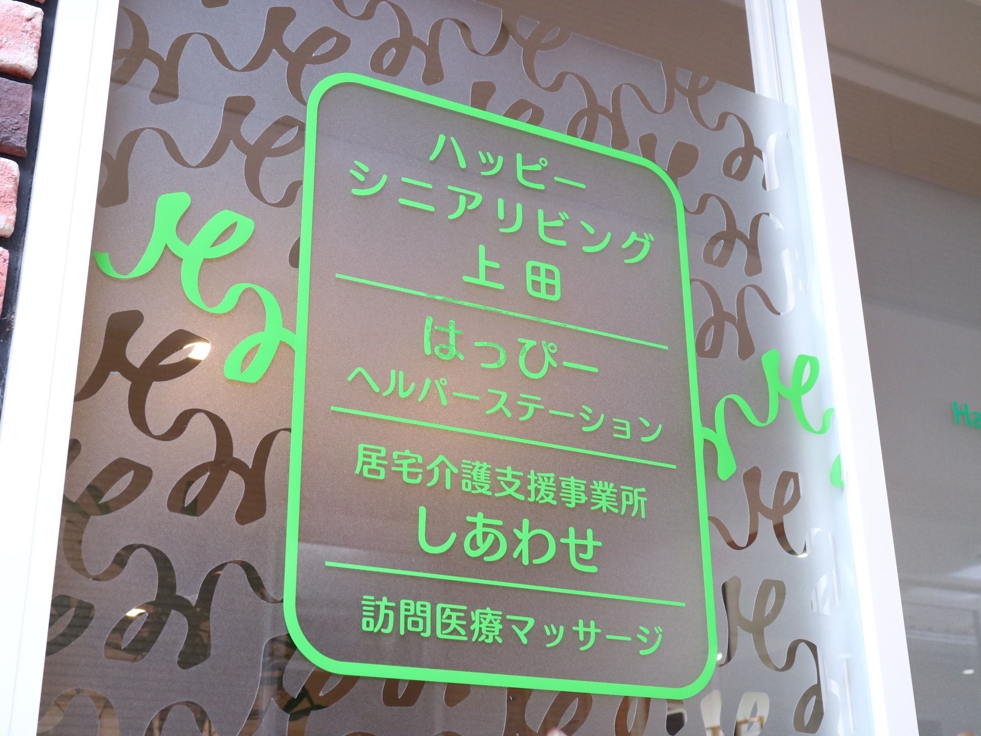 エントランスのガラス戸に、建物内に入っている施設の名称が記載されている。施設のロゴマークに合わせて、リボンが一緒に描かれている。
