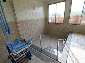 大きな埋め込み式の浴槽が設置された浴室。そのそばにはブルーの車いすのようなリフトがあり、天井からひもがぶら下がっている。