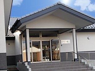 ダークグレーの瓦屋根が付いた白壁の建物。玄関には屋根があり、手すり付きの階段が設置されている。