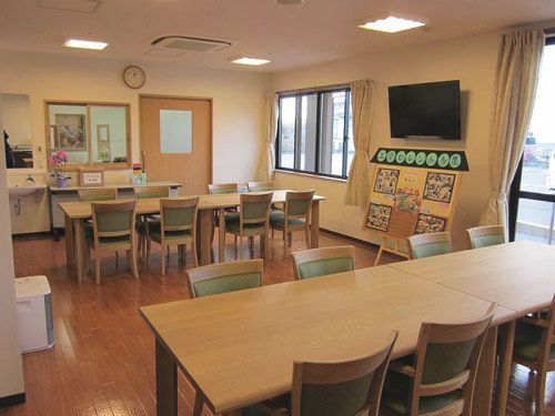 薄い色の木目のダイニングテーブルと椅子が数脚置いてある。壁に洗面台と時計、薄型テレビが設置されている。