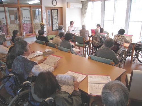 大きな窓から光が入る明るい部屋で、大勢の入居者様が椅子や車椅子に腰を掛けて紙面に目を通している。壁にエアコンと時計が設置されている。
