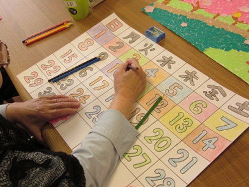 眼鏡をかけた人物が、曜日と日付がカラフルに書かれたカレンダーを作成している。机の上に鉛筆や消しゴムが置いてある。