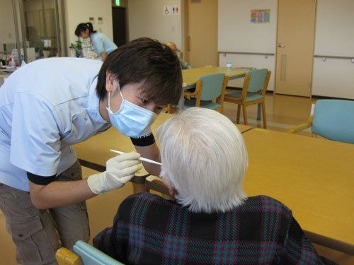 茶色いテーブルの前に置いてある緑色の椅子に、白髪の女性が腰を掛けている。マスクを着用した男性が手袋をはめている。