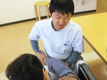 クリーム色の床の上に、丸い木材の椅子が置いてある。白衣を着用した男性が年配の女性の足をマッサージしている。