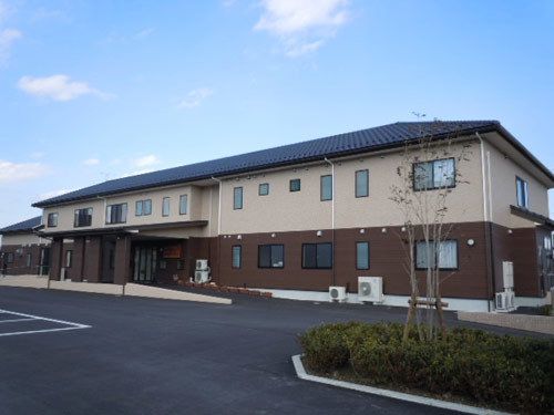 青空の下に、茶色とベージュ色の２階建ての建物が建っている。植栽スペースに樹木が植えられている。エアコンの室外機が数台置いてある。