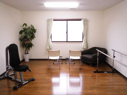 白いクロスの壁とフローリングの床に腰高窓が設置された明るい部屋である。観葉植物が飾られ、平行棒や健康器具が置いてある。