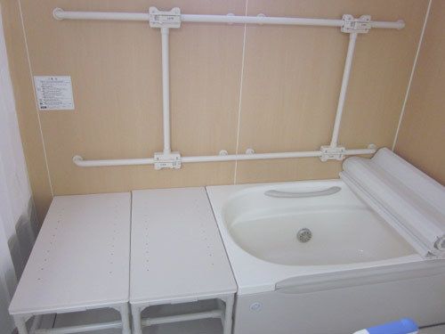 ベージュ色の壁には縦横方向の手すりが設置されている。浴槽にシャッタータイプの蓋が半開きの状態で置いてある。