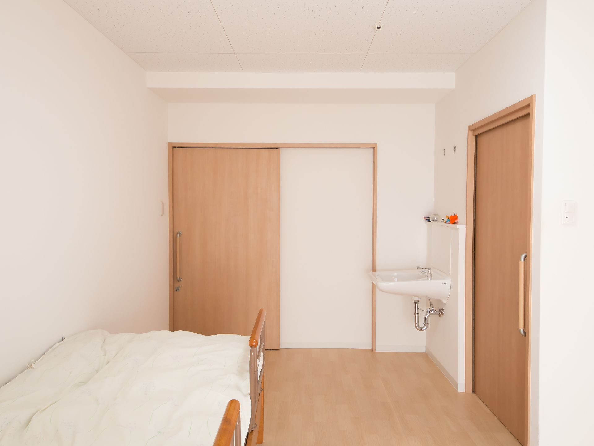 ナチュラルカラーのフローリングの床と、同色の扉のある部屋に、介護ベッドが１台置かれている。部屋の角に、広いシンクの洗面台が設置されている。