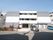 ３階建てで、グレーと白色の壁の建物で、手前右側に駐車スペースがある。左側に木の植えられたスペースが設けられている。