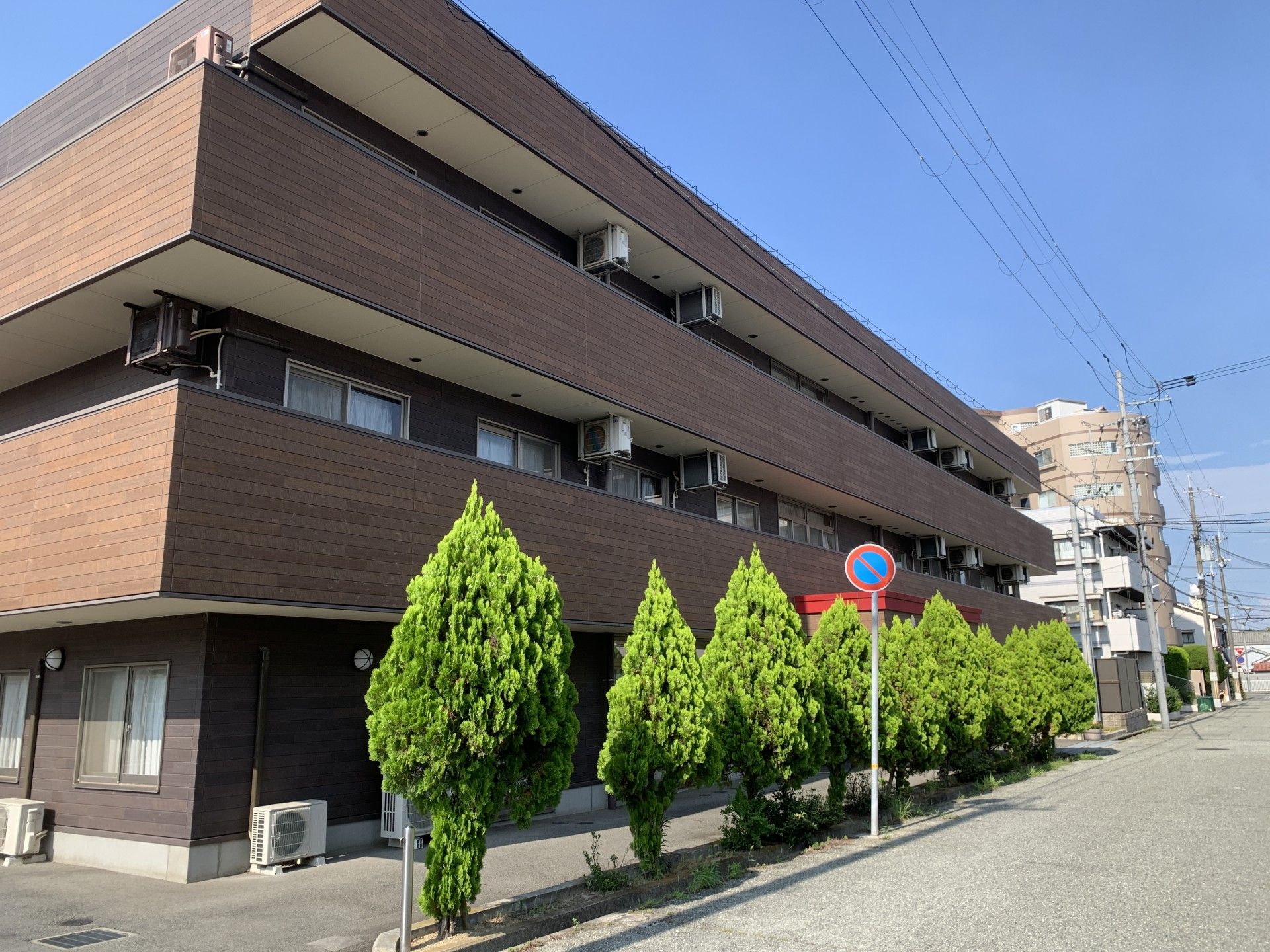 3階建ての建物はブラウン系で各フロアにベランダがあり、緑に囲まれている。玄関の屋根にはホーム名が書かれ、日影ができている。