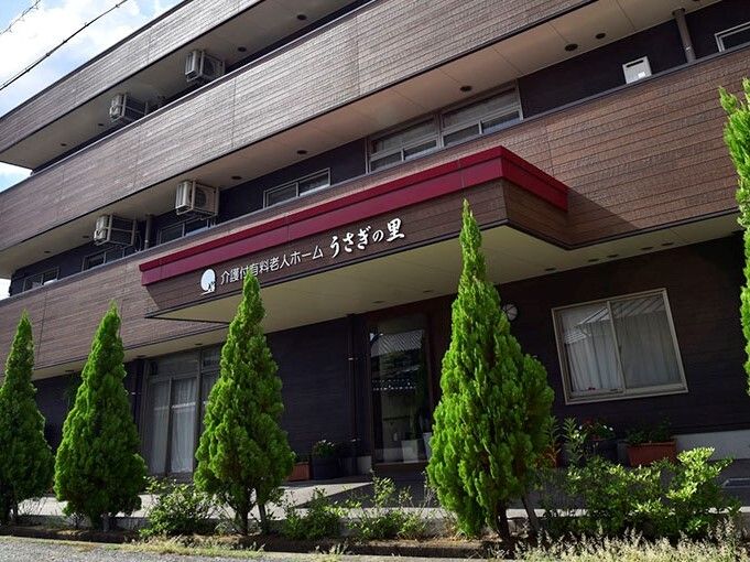 ブラウン系の3階建ての建物は緑に囲まれていて、各フロアにベランダがあり、玄関の屋根にはホーム名が書かれている。