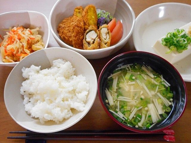 木製のテーブルの上には白いご飯とみそ汁、野菜のフライの盛り合わせ、煮物、冷ややっこなどが並んでいる。