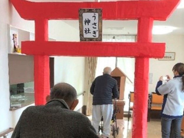 館内に設置された赤い鳥居と社は手作りで、男性入居者が2名お参りしている姿を女性スタッフがカメラに収めている。
