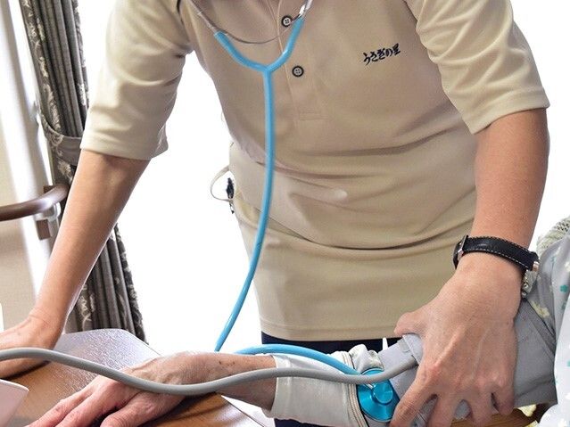 聴診器を持ったスタッフのユニフォームにはホーム名が書かれている。細く筋張った入居者の腕には血圧測定機がセットされている。