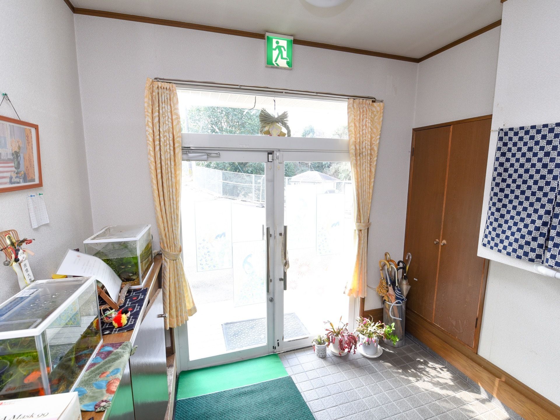 ガラス張りの玄関は、石畳の上にマットが敷かれ、サボテンなどの鉢植えがいくつか並んでいる。壁際には棚の上に水槽があり、金魚が泳いでいる。