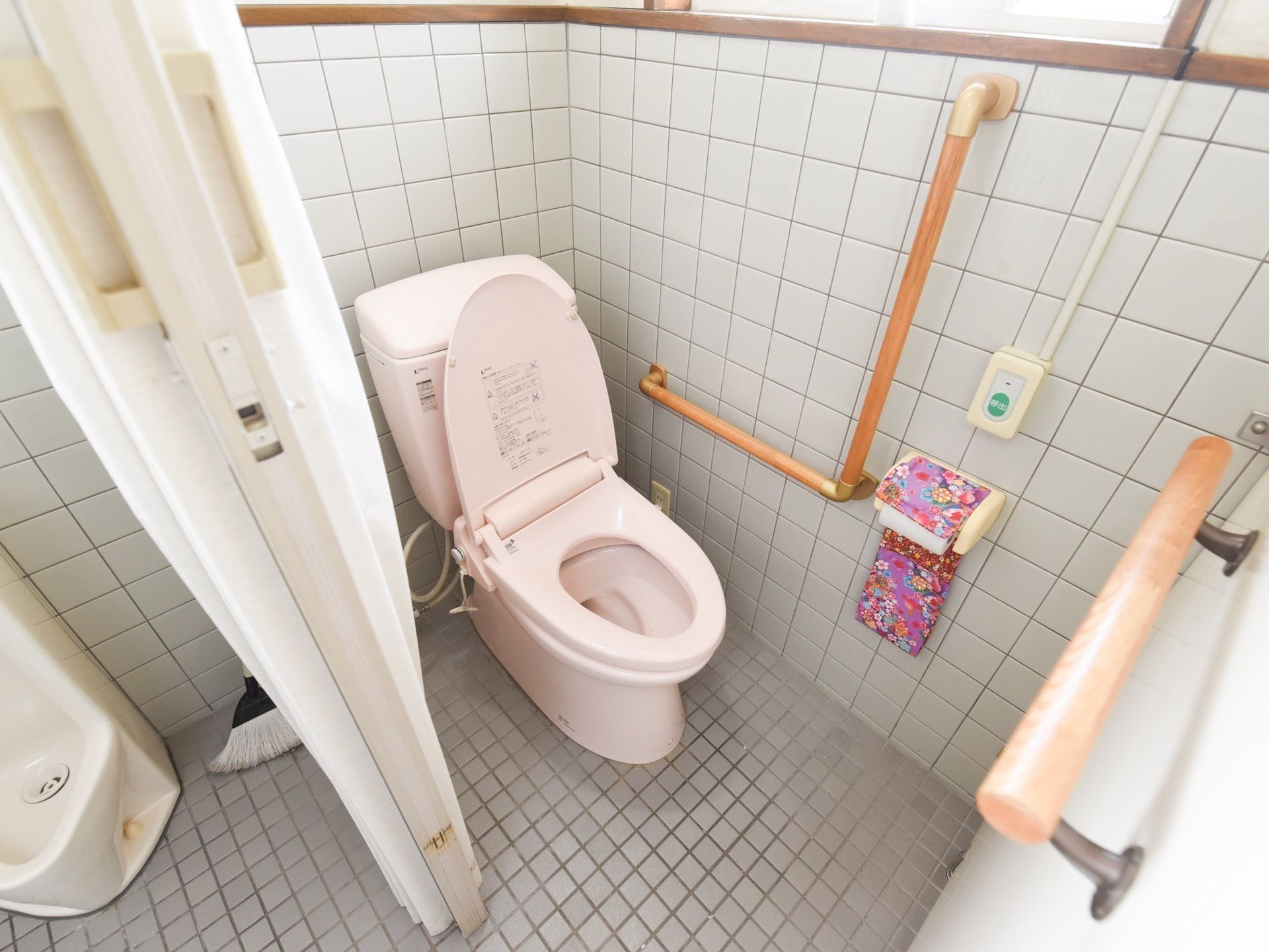 トイレには、タイル張りの壁面にＬ字型の手すりとナースコールが取りつけられている。仕切りを隔てた隣には、男性用便座が設置されている。