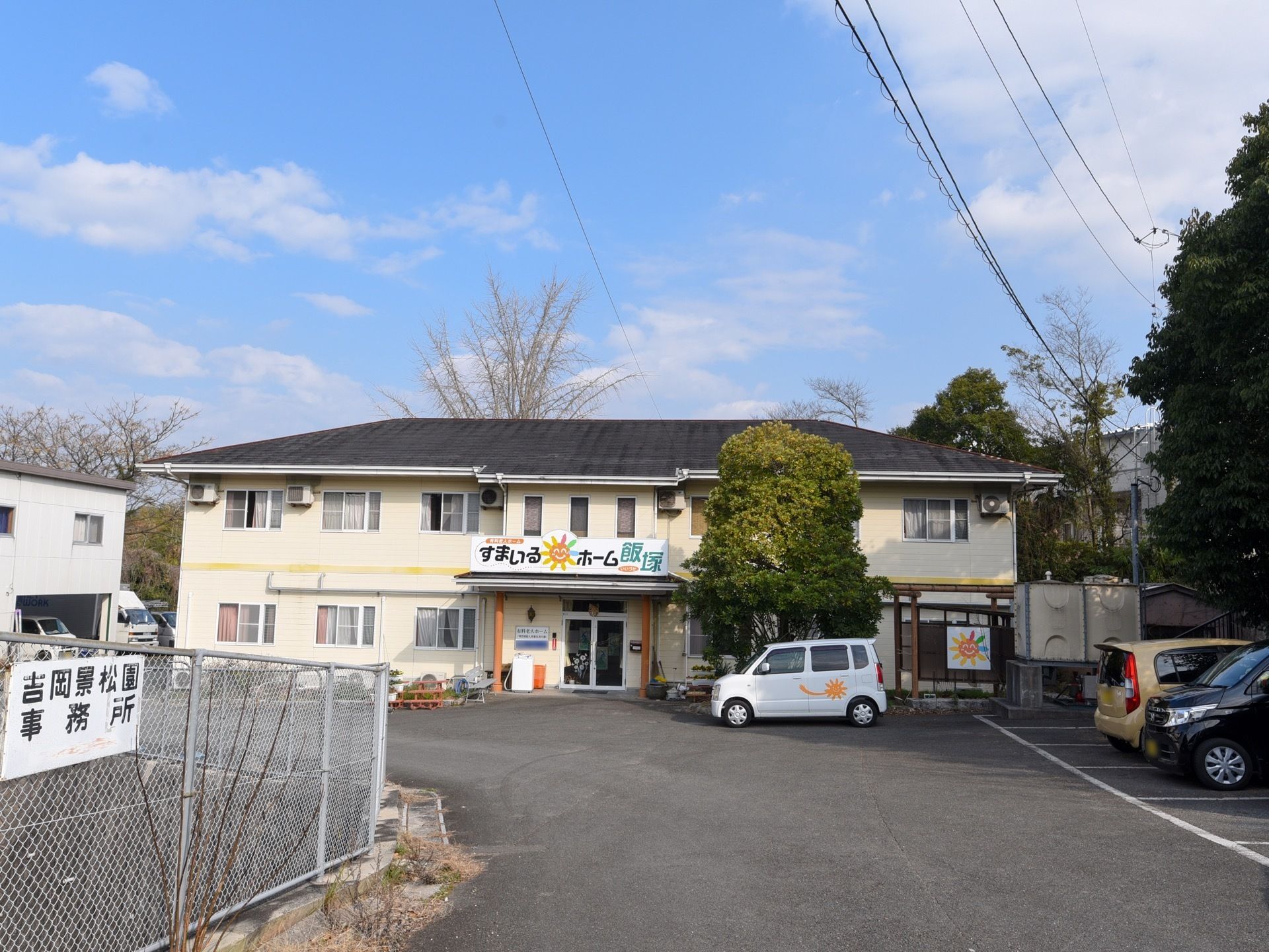 白色を基調とした外壁の２階建ての施設である。広い敷地内に多くの駐車スペースが確保され、施設のマークがついた車も停まっている。