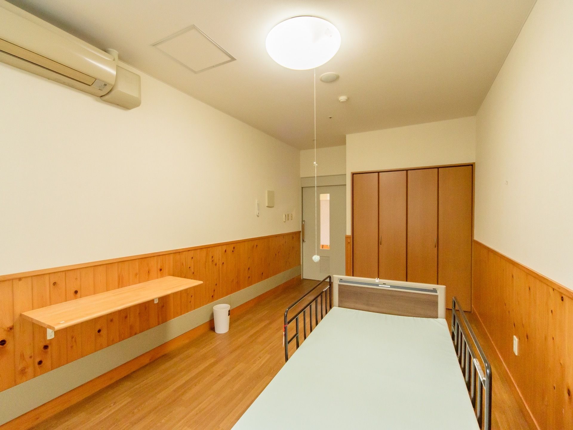 木のぬくもりが感じられる温かみのある居室で、壁には机が取り付けられている。天井には、紐付きの照明が設置されている。