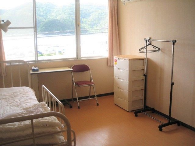 右手にはハンガーラックや収納ケース、左手にはベッド、窓際にはパイプいすが置かれた居室。
