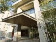 大きな車寄せが玄関に設置されているベージュの外壁の大きな建物。周りには草木が植えられている。