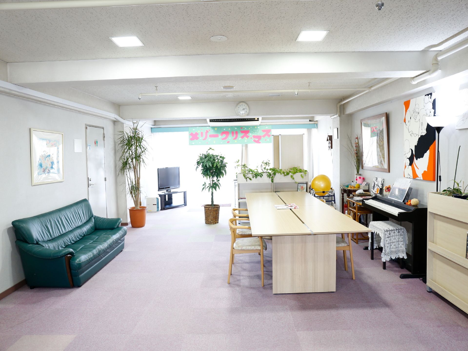 壁、天井、床とも白系の色である。壁際に長がけソファがある。反対側の壁に電子ピアノ、部屋の角にテレビがある。長いテーブルと肘掛け椅子がある。