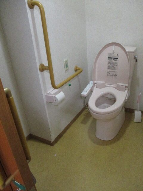 薄いピンクの便座が付いたトイレのそばにはエル字型の手すりやグリーンの呼び出しボタンを設置している。