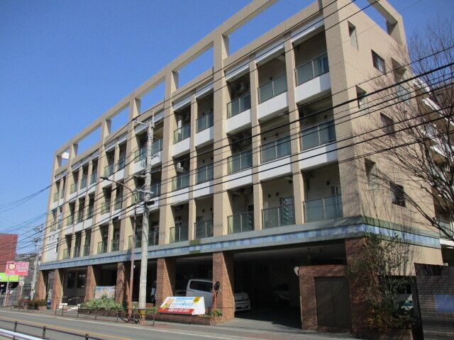 ベージュの外壁の４階建ての建物の周りには草木が得られ、各フロアにベランダがある。