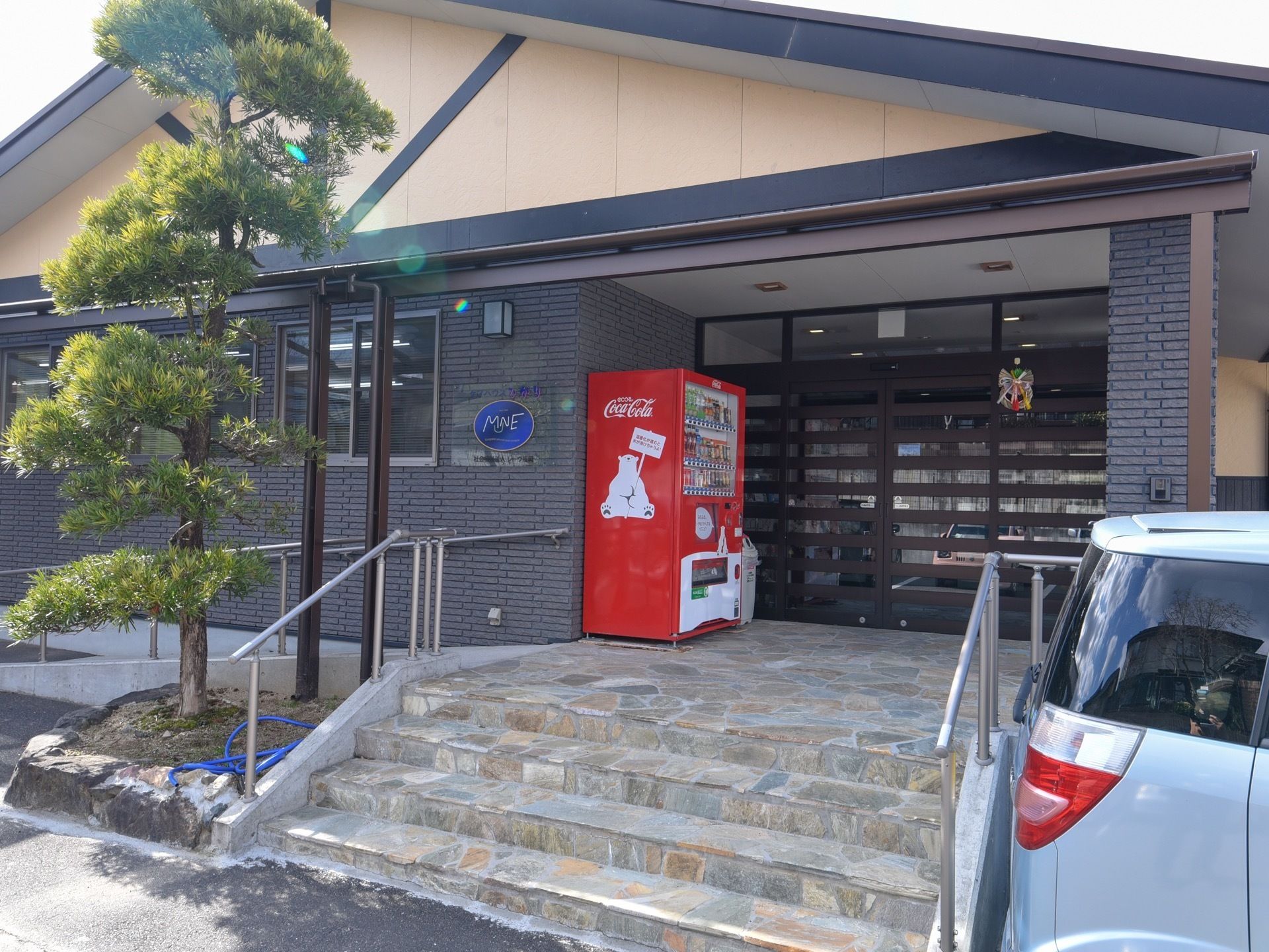 幅の広い階段があり、両側には手すりが取りつけられている。植え込みには1本の木がある。自動販売機が設置されている。