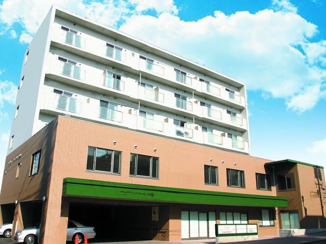 6階建ての建物はベージュと白の2色使いの外壁で、窓がたくさん付き、1階部分の駐車場には車も止まっている。