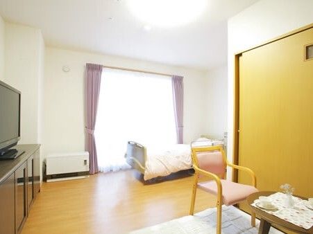 レースカーテンとカーテンが付いた大きな窓のそばには寝具付きのベッドが置かれ、タンスの上にはテレビがあり、テーブルセットも置かれている。