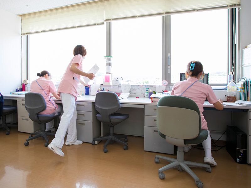 ピンクのユニフォームを着た女性スタッフが３名、机に向かって作業をしている。一人は立っている。