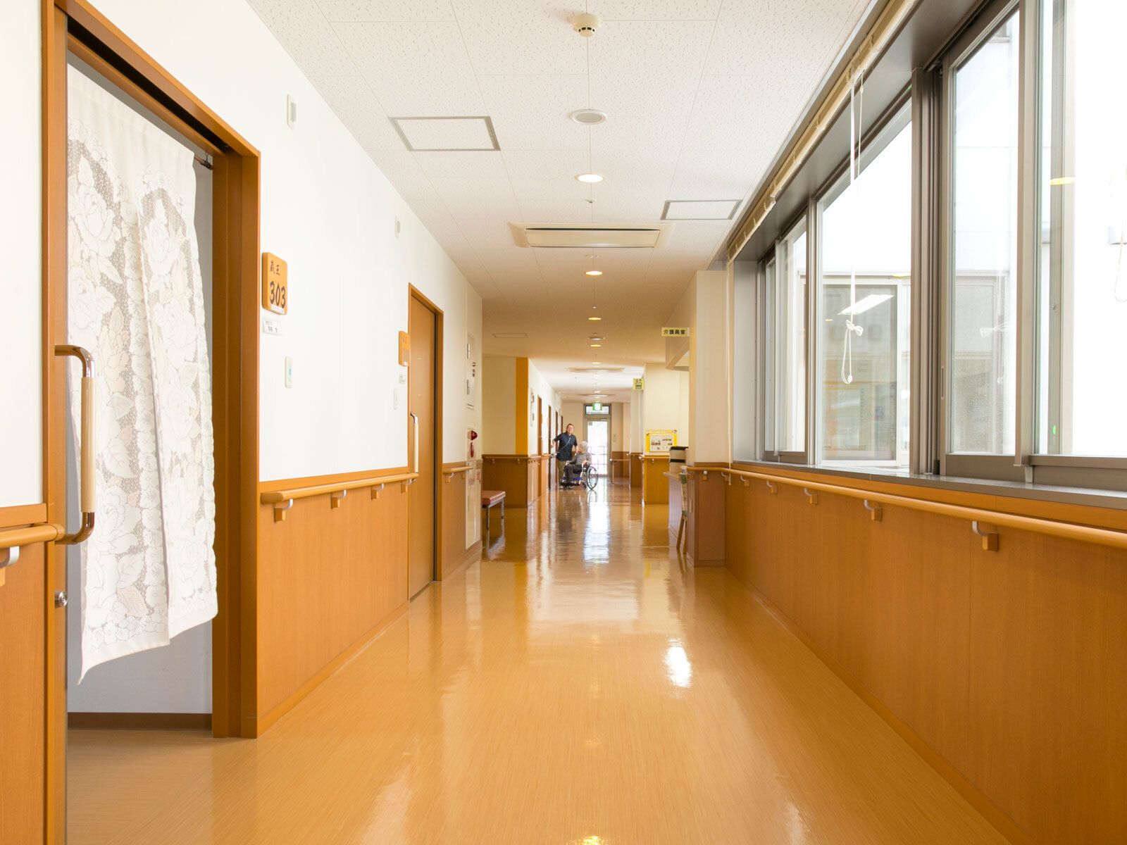 手すりが両サイドについたまっすぐな廊下。左側には暖簾がかかった部屋があり、通路には何も置かれていない。