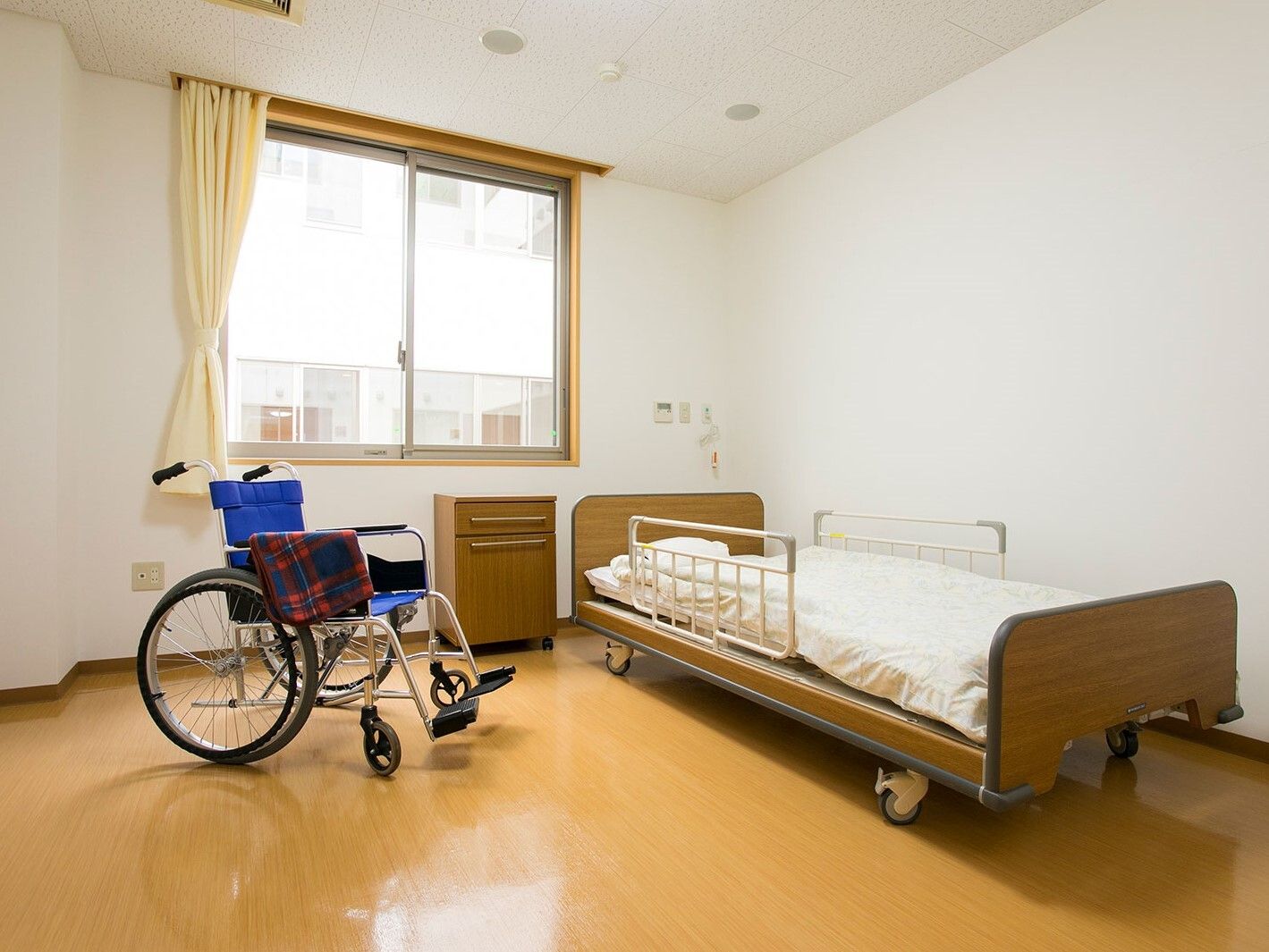 寝具付きのベッドが置かれたフローリングの部屋には車いすが置かれ、大きな窓もある。