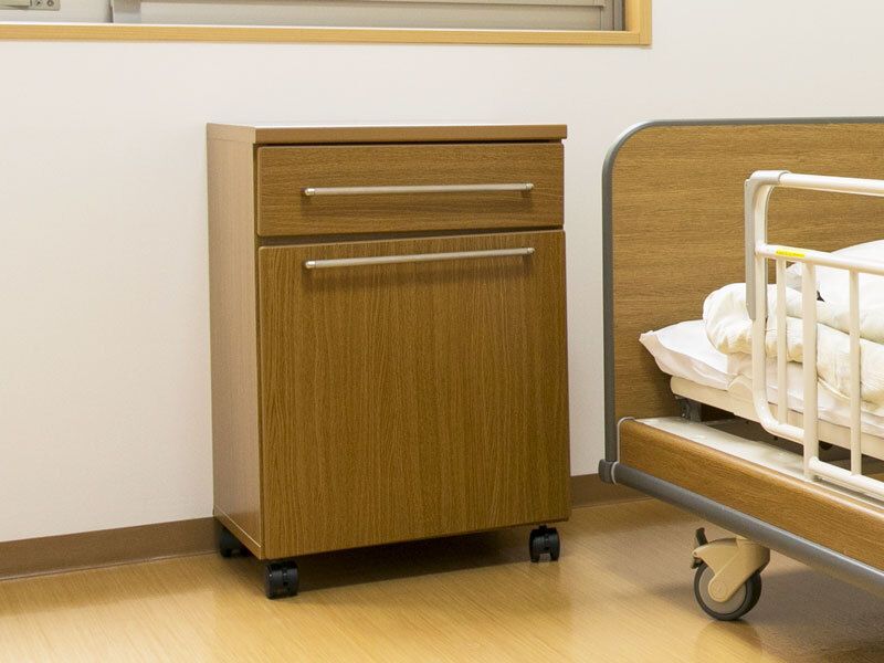 寝具付きの介護ベッドには手すりが付いている。そのそばには引き出し付きの小さな収納家具がある。