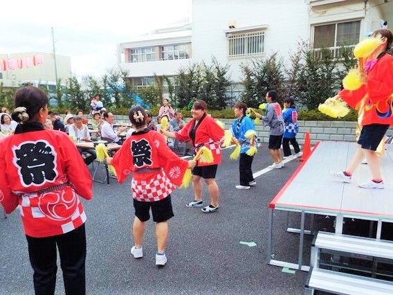 椅子に座って集合している入居者様の前で、多数のスタッフがお揃いのはっぴを着て踊っている屋外の様子