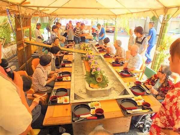 野外の屋根付きテントの中で、大勢の入居者様が並んで着席され、流しそうめんの食事会を催している様子
