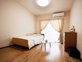 フローリングの床の小さなワンルーム。タンス、ベッド、奥のほうに窓が写っている